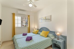 Un dormitorio con dos camas, un ventilador de techo, una ventana con cortinas, un cuadro en la pared y una mesita de noche con lámpara.