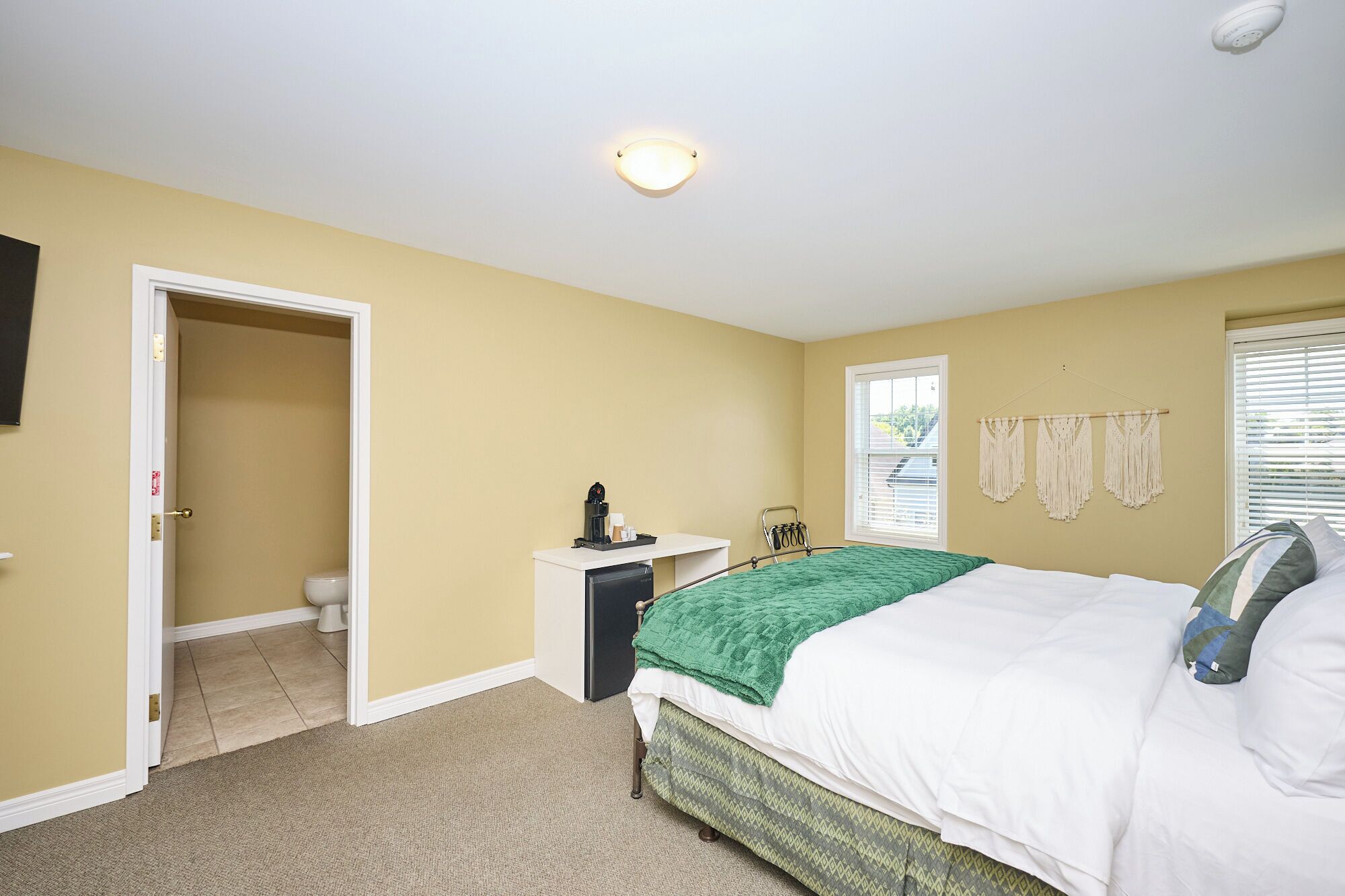A bedroom with a bed, a bedside table, a window with blinds, a door leading to a bathroom, and a ceiling light.