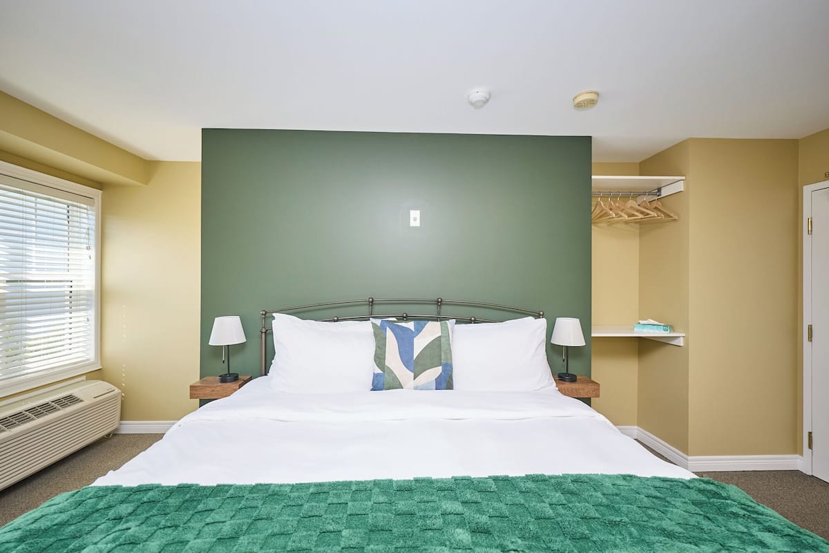Een netjes opgemaakt bed met witte lakens en een groene plaid, een nachtkastje met een lamp, en een raam met jaloezieën.