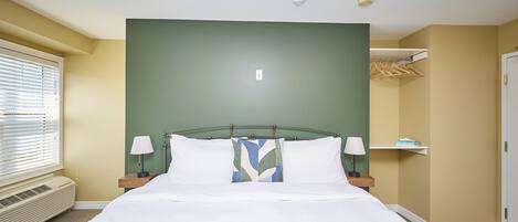 Una cama bien hecha con sábanas blancas y un plaid verde, una mesita de noche con lámpara, y una ventana con persianas.