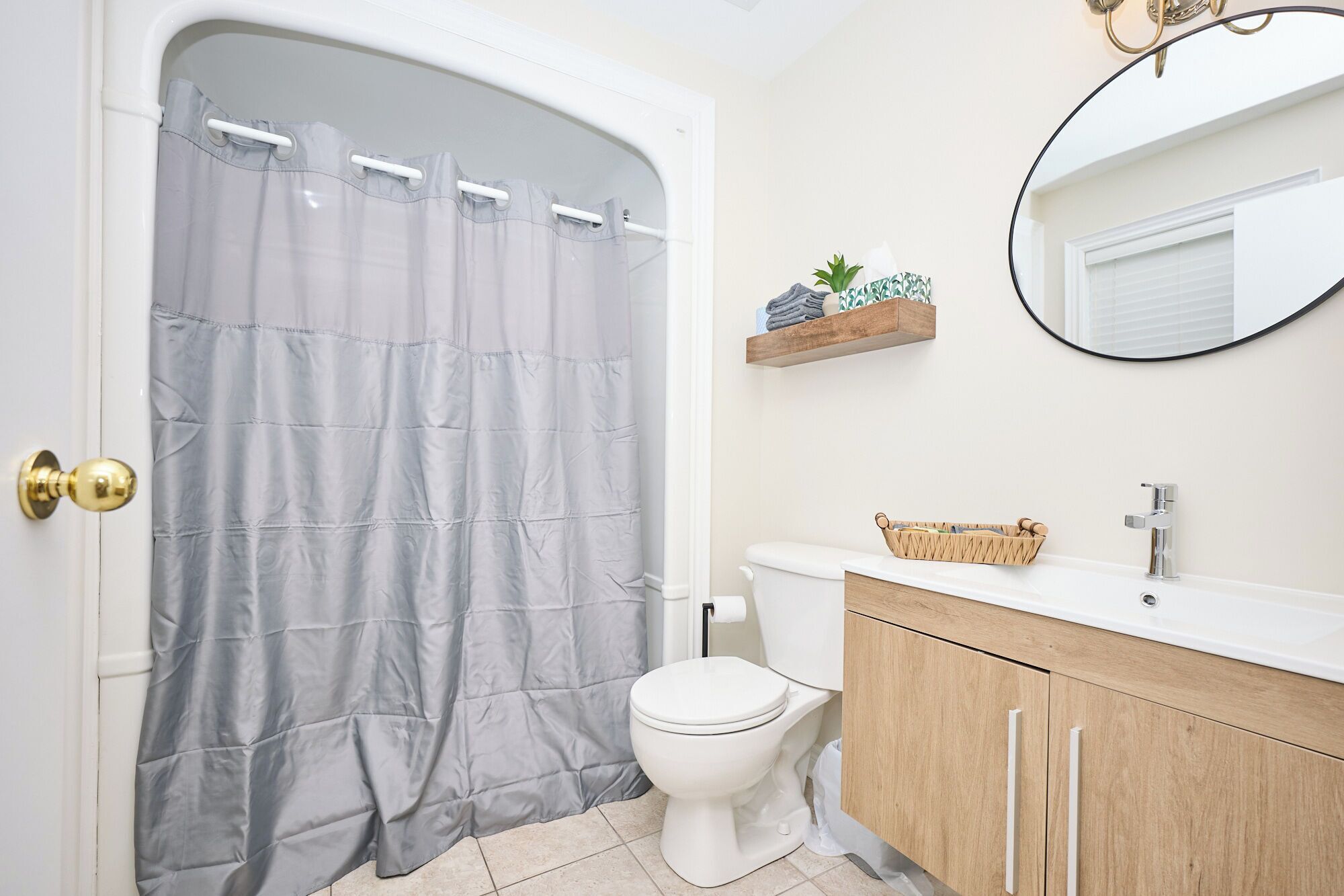 A bathroom with a shower curtain, toilet, vanity with a mirror, and a shelf with toiletries.