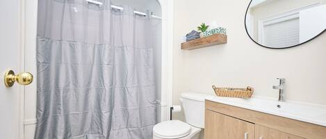 A bathroom with a shower curtain, toilet, vanity with a mirror, and a shelf with toiletries.