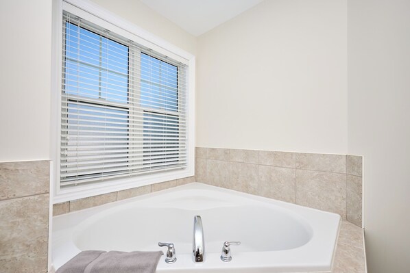 A bathroom with a large bathtub, a window with blinds, and tiled walls.