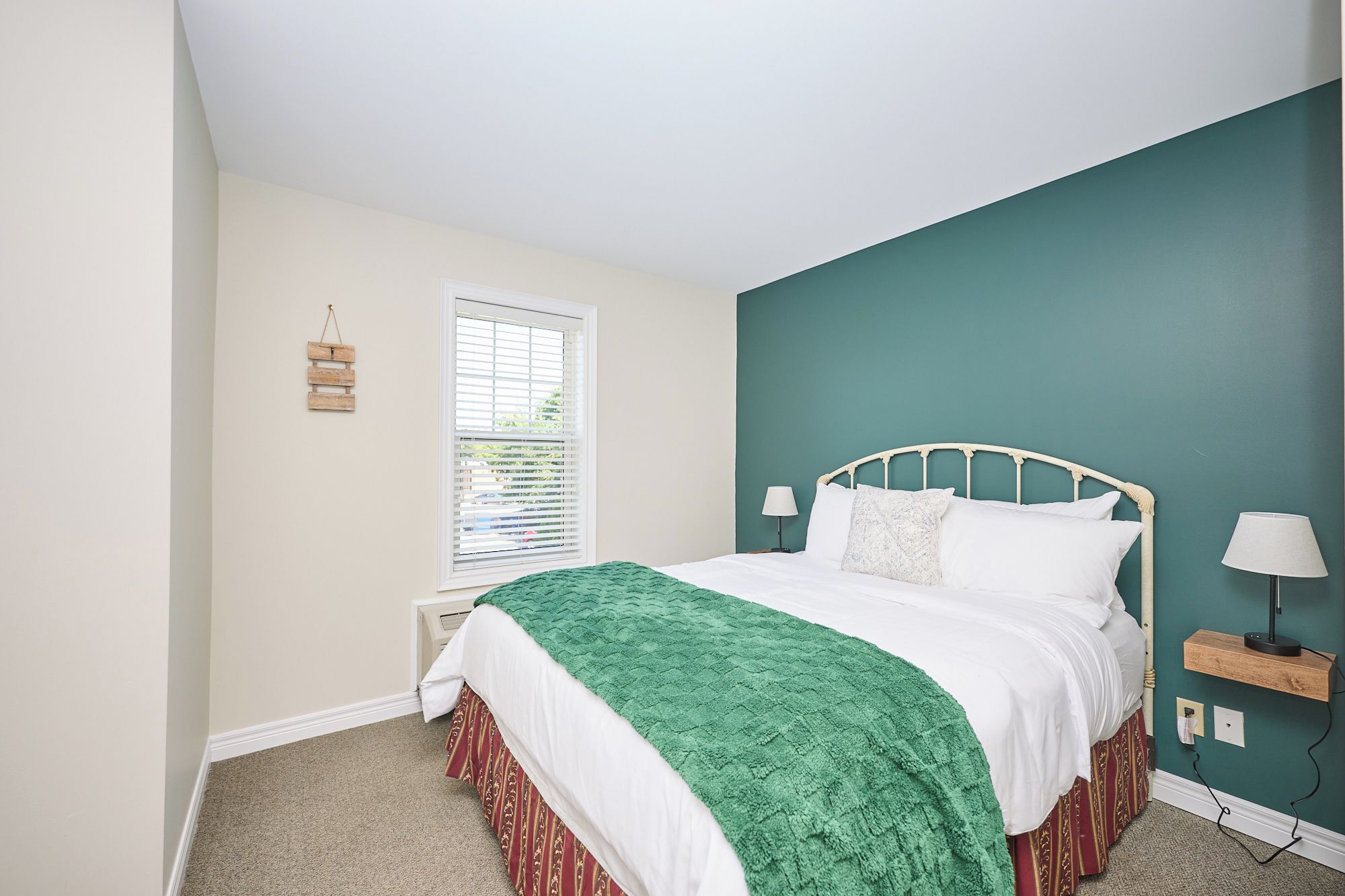 A bedroom with a bed, a window with blinds, a bedside table with a lamp, and a wall-mounted shelf.