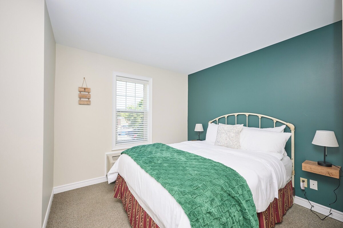 A bedroom with a bed, a window with blinds, a bedside table with a lamp, and a wall-mounted shelf.