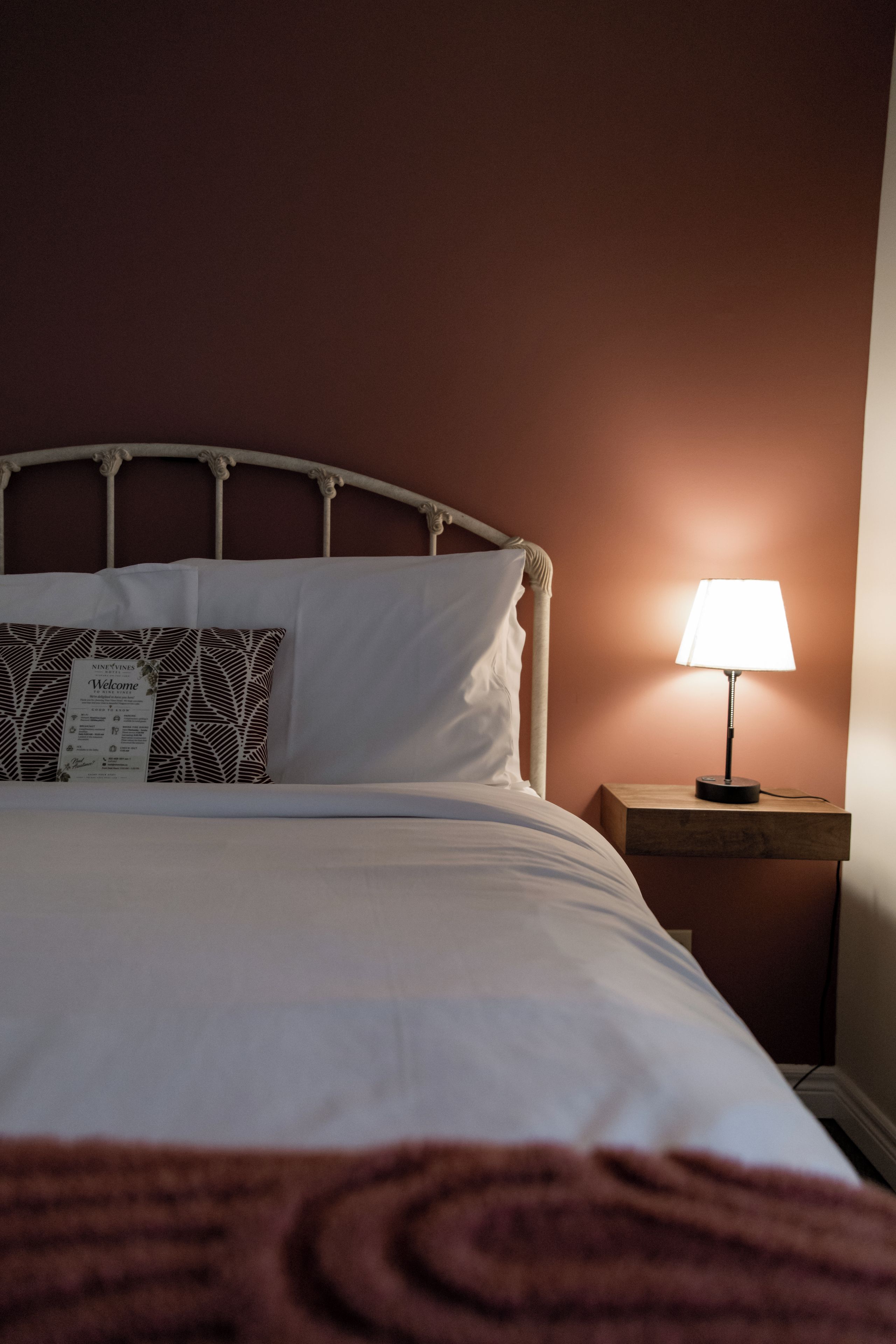 A neatly made bed with white bedding and a patterned pillow, a bedside lamp, and a wooden nightstand.