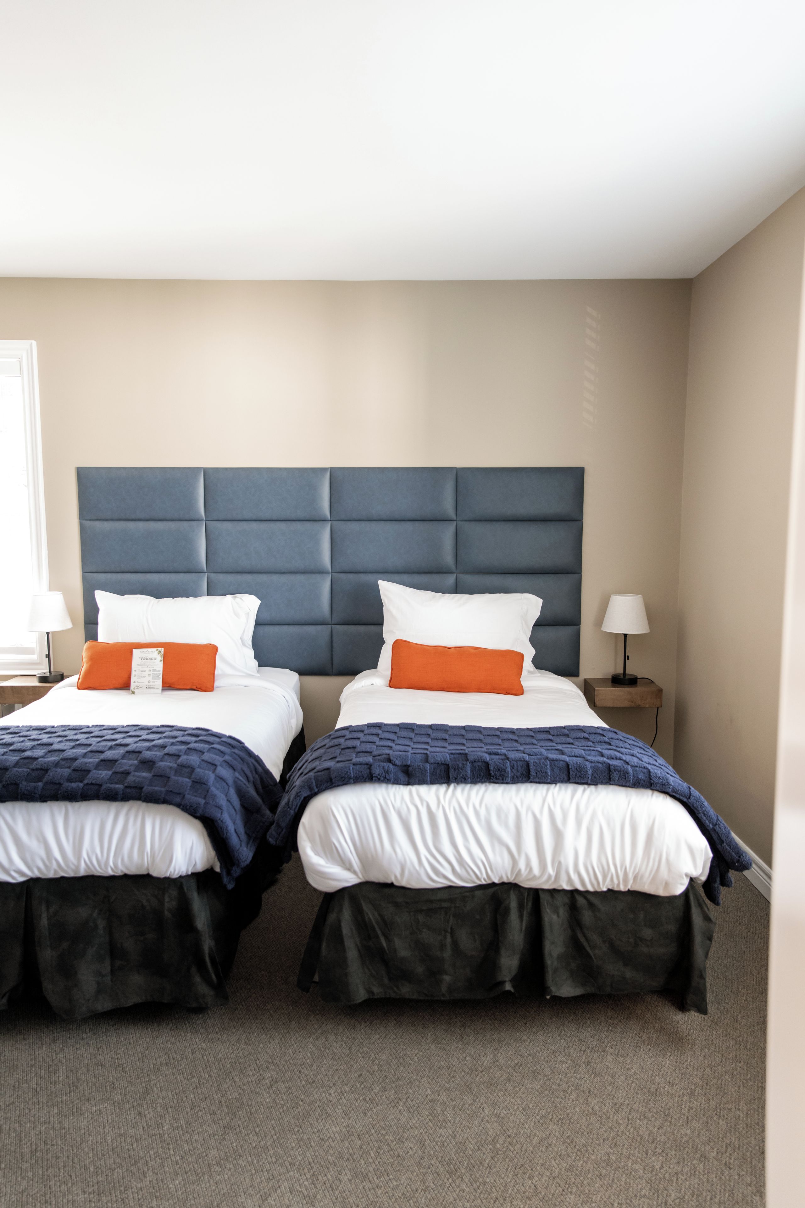 Une chambre d’hôtel avec deux lits, chacun doté d’une tête de lit, de draps blancs et d’oreillers orange. Il y a une table de chevet avec une lampe de chaque côté des lits.