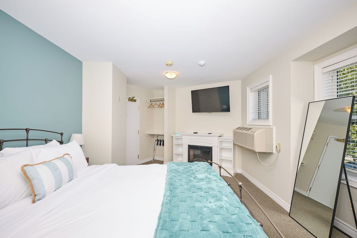 Een slaapkamer met een bed, een televisie boven een open haard, een spiegel en een raam met zonwering.