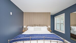 A bedroom with a bed, a window with blinds, a wall-mounted light, and a mirror.
