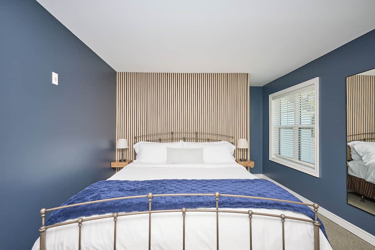 Een slaapkamer met een bed, een raam met jaloezieën, een wandlamp en een spiegel.
