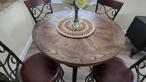 Ein runder Holztisch mit einer gewebten Tischmatte, umgeben von vier gepolsterten Stühlen mit Metallgestellen. Auf dem Tisch steht eine Vase mit Blumen.