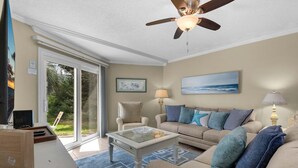 Ein Wohnzimmer mit Glastisch, beiger Couch mit blauen und grauen Kissen, Deckenventilator und einem großen Fenster mit Blick ins Grüne.