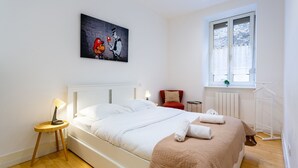 A bedroom with a bed, a red chair, a small table with a lamp, a painting on the wall, and a window with a view.