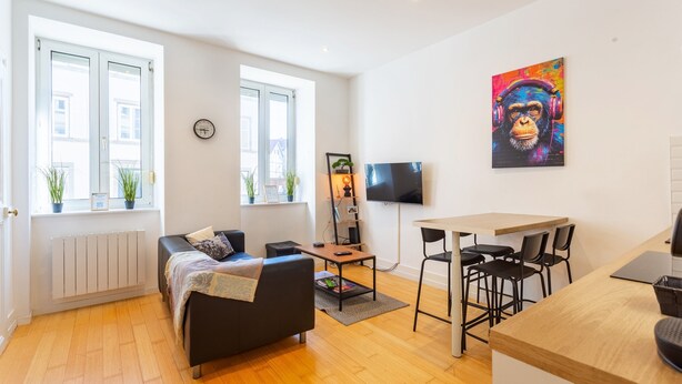A modern living room with a sofa, a coffee table, and a dining area with chairs. There is a TV mounted on the wall and a window with plants on the sill.