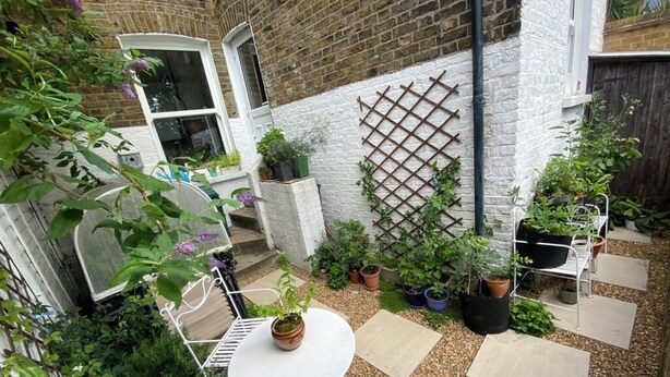 Un pequeño jardín resguardado con pared de ladrillo blanco, celosía, plantas en macetas y una mesa redonda con sillas.