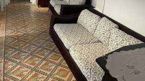 Une pièce avec un tapis à motifs, un canapé recouvert d’un tissu à fleurs, une armoire en bois et un lit avec un oreiller vert.