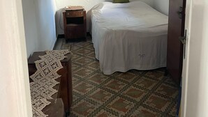 Une petite pièce étroite, avec un lit simple, une table de chevet, une armoire en bois et un sol à motifs.