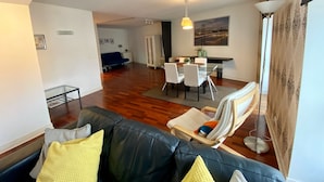 A spacious living area with a black leather sofa, a glass dining table with white chairs, a blue sofa in the background, and a painting on the wall. - Downtown River Side by Homing (Lisbon)