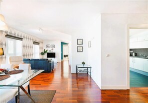 A modern living room with a glass dining table, a sofa, and a TV. There is a kitchen visible through an open door.