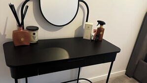 A vanity table with a round mirror, a small vase with black sticks, a vanilla candle, and a few skincare products.