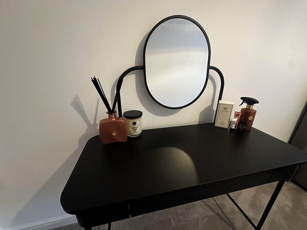 Een zwarte tafel met een ronde spiegel, een kleine vaas met zwarte takken, een geurkaars met vanilleboon en enkele persoonlijke verzorgingsproducten.