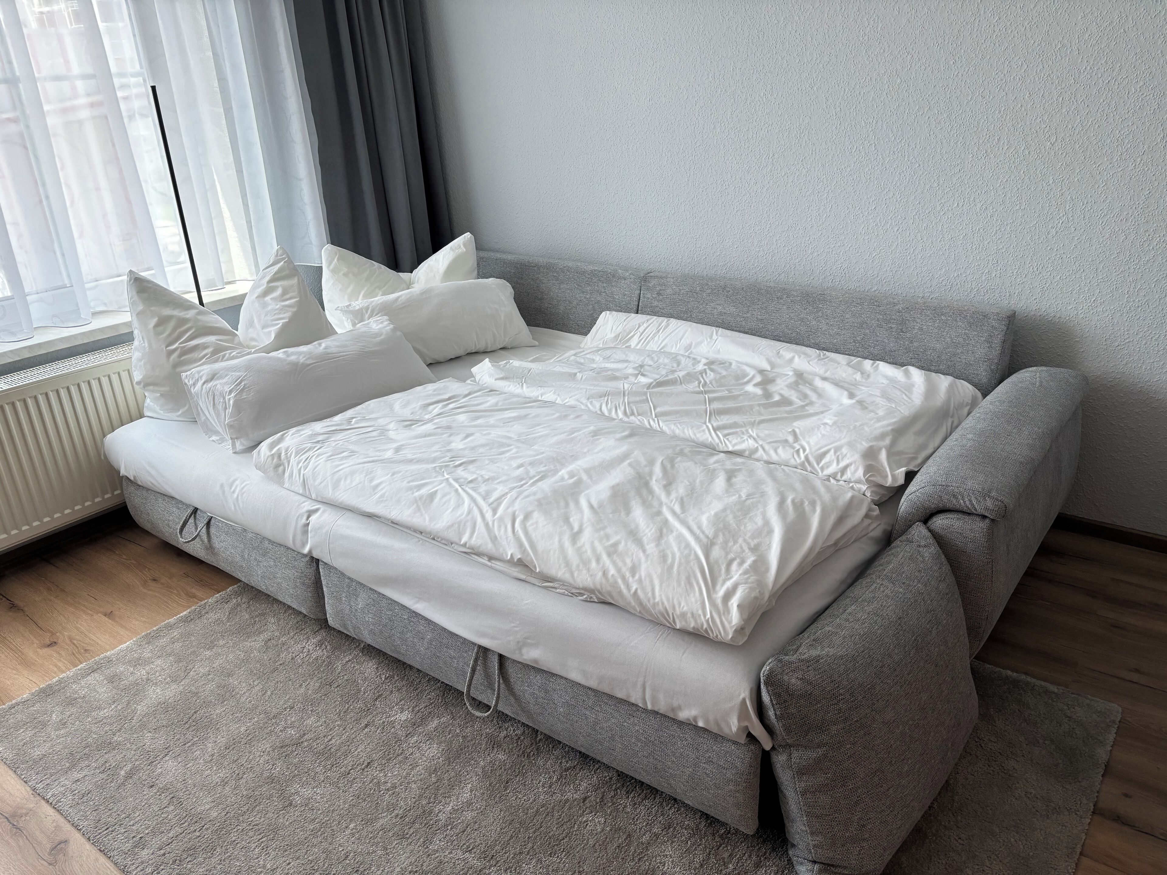 A neatly made bed with white bedding and pillows, a grey sofa, and a grey rug in a room with a radiator and a window with curtains.