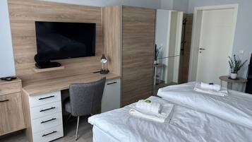 Une chambre d’hôtel avec un bureau en bois, une télévision, une chaise, un lit avec du linge de lit blanc et une petite table sur laquelle est posée une plante.