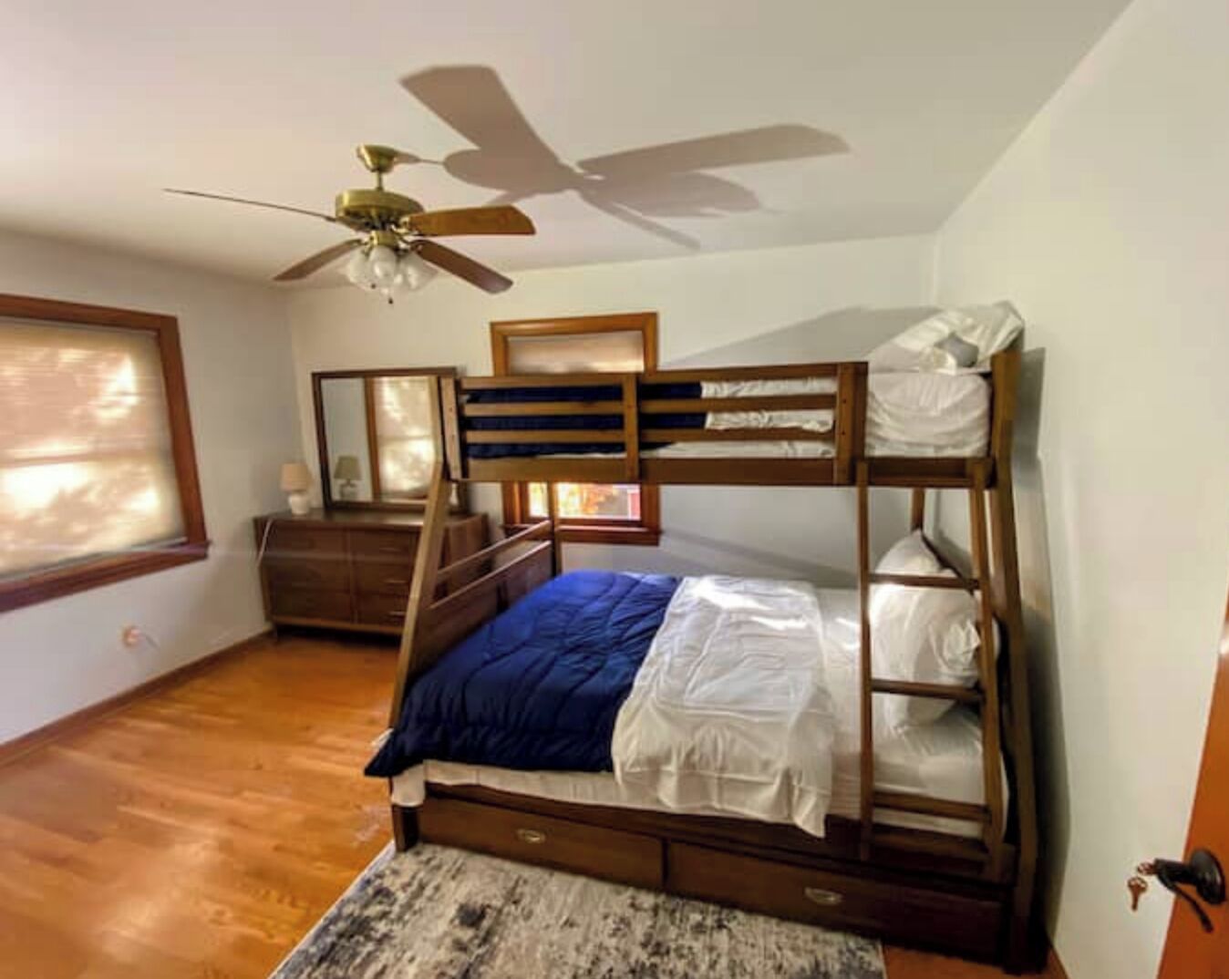 Dormitorio con literas, ventilador de techo, piso de madera, cómoda y ventana con persianas.