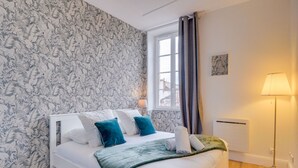 Ein ordentlich bezogenes Bett mit weißen und türkisblauen Kissen, einer grünen Tagesdecke und einem Fenster mit Vorhängen.