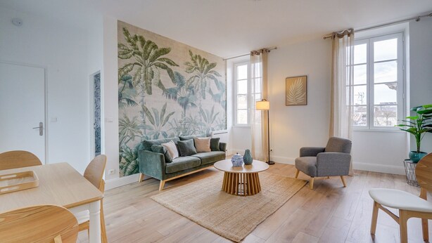 Ein Wohnzimmer mit Holzboden, einem grünen Sofa, einem grauen Sessel, einem runden Couchtisch und einem großen Wandgemälde mit tropischem Motiv.