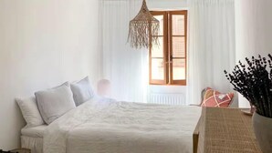Un dormitorio con cama, lámpara colgante, ventana con cortinas y una silla.