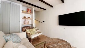 Una sala de estar con sofá, una silla de mimbre, una mesa redonda y un televisor montado en la pared.