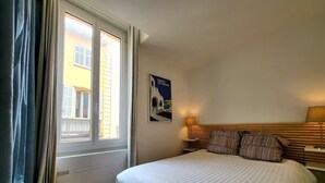 A hotel room with a bed, a nightstand, a lamp, a poster, and a window with curtains. - L'Aiguillon Antibois - Proche Port (Antibes)
