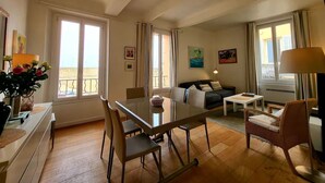 A spacious living room with a glass dining table, comfortable seating, and large windows offering a view. - L'Aiguillon Antibois - Proche Port (Antibes)