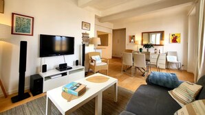 A modern living room with a flat-screen TV, a white coffee table, a grey sofa, and a dining area with white chairs and a table. - L'Aiguillon Antibois - Proche Port (Antibes)