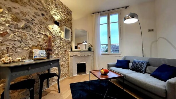 Un salón acogedor con pared de piedra, chimenea, sofá con cojines azules, escritorio con silla y lámpara de pie.