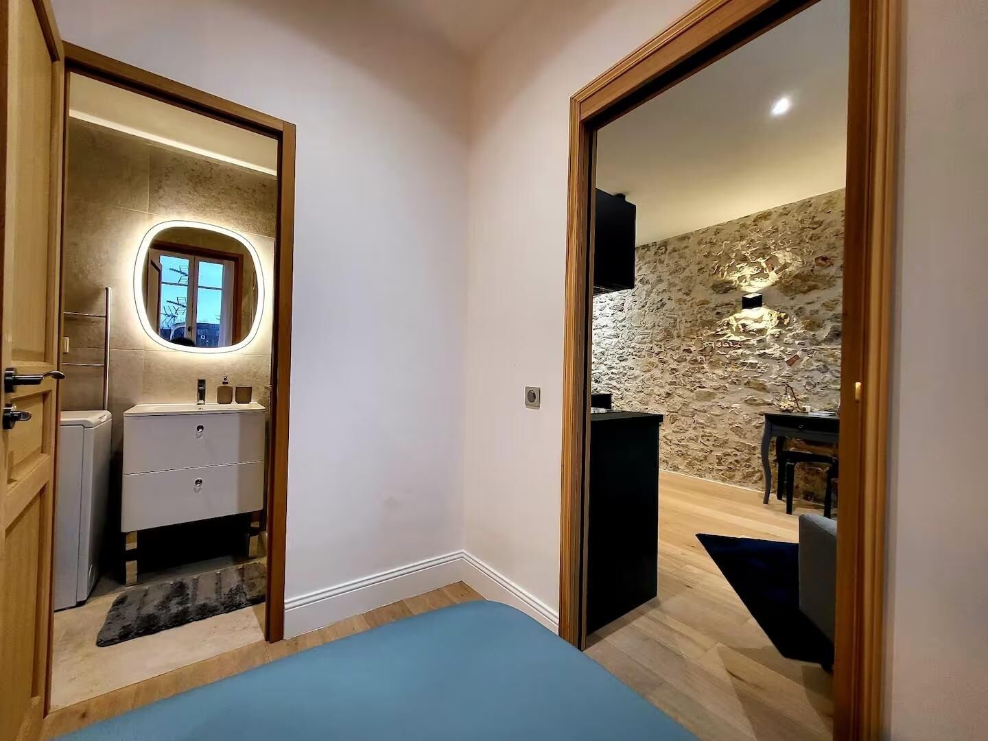 Een slaapkamer met een bed, een dressoir en een spiegel. Er is een stenen muur en een raam in de aangrenzende kamer.
