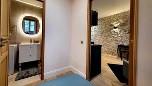 Un dormitorio con una cama, un aparador y un espejo. Se observa una pared de piedra y una ventana en la habitación contigua.