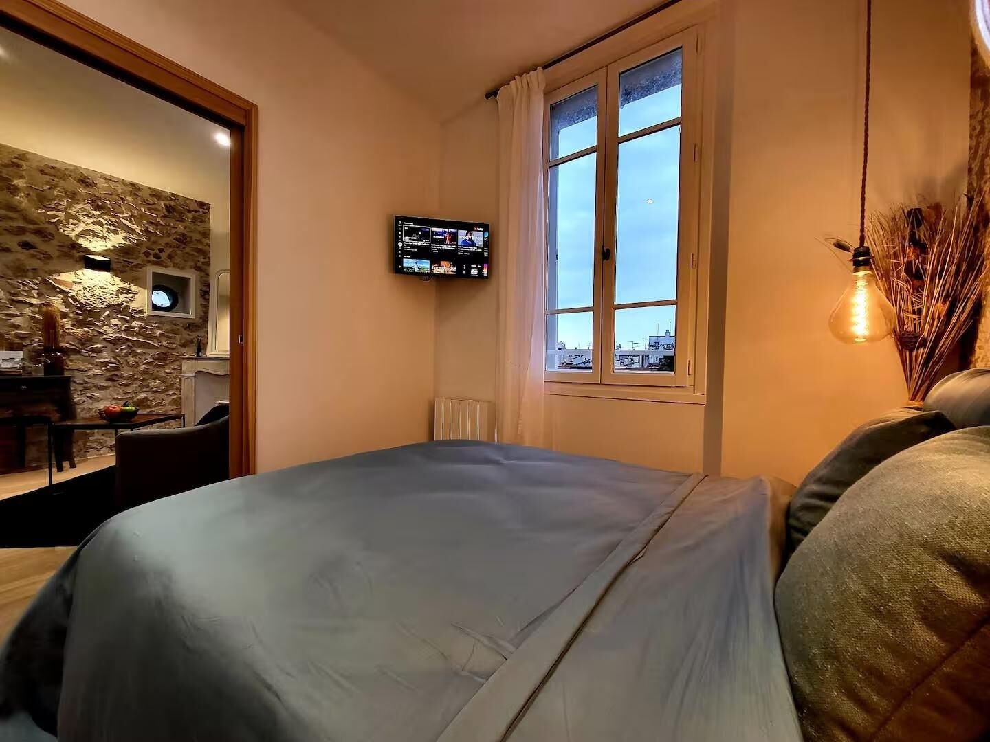 A bedroom with a bed, a TV mounted on the wall, a window with a view of buildings, and a stone wall in the background.