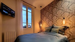 Un dormitorio con cama, televisor montado en la pared, ventana y papel tapiz estampado.