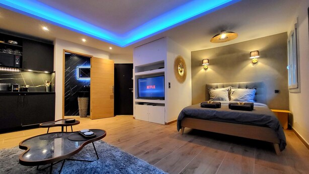 Une chambre moderne avec un lit, une télévision et un coin cuisine visible à travers une porte ouverte.