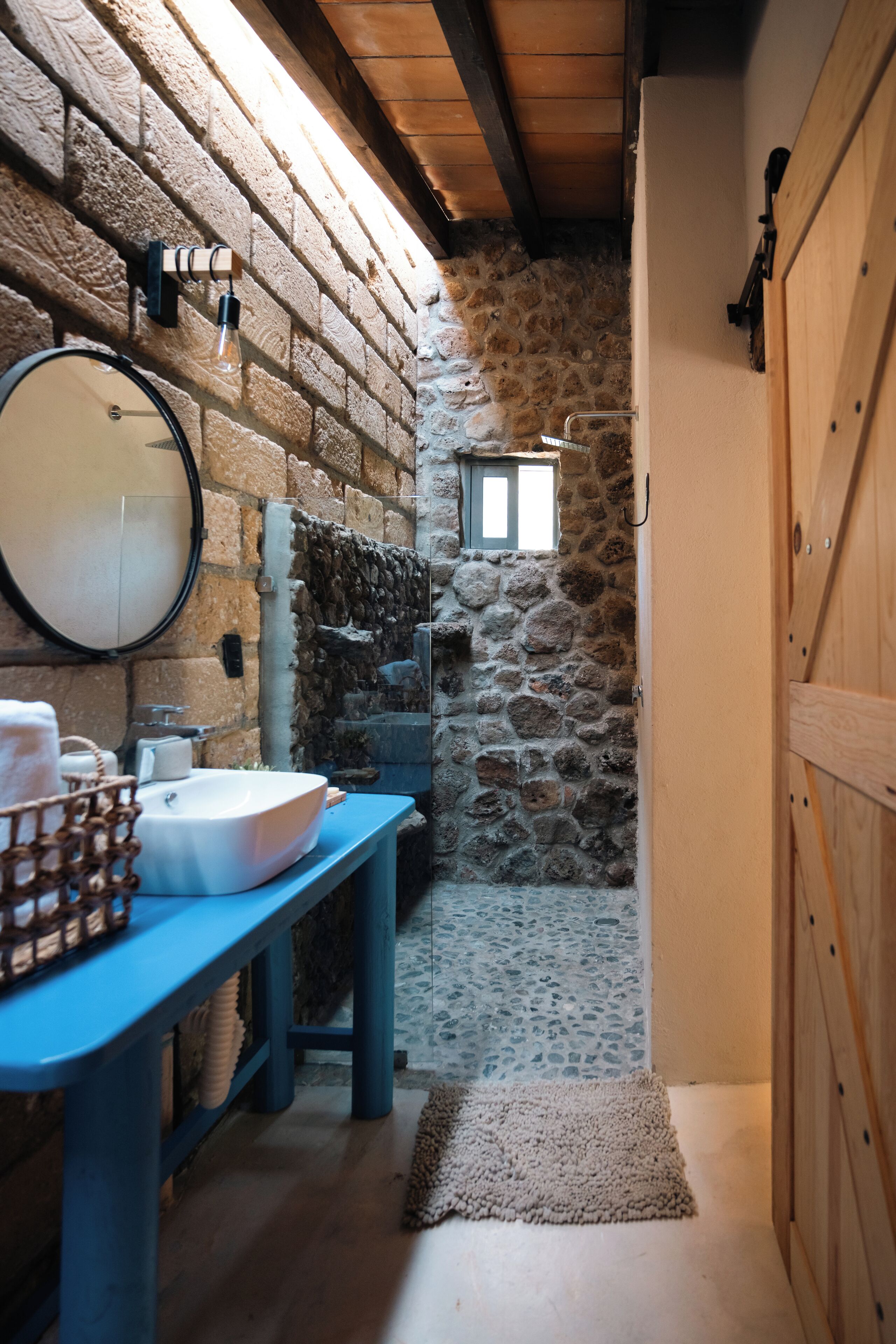 A narrow stone-walled bathroom with a wooden bench, a sink, and a mirror.