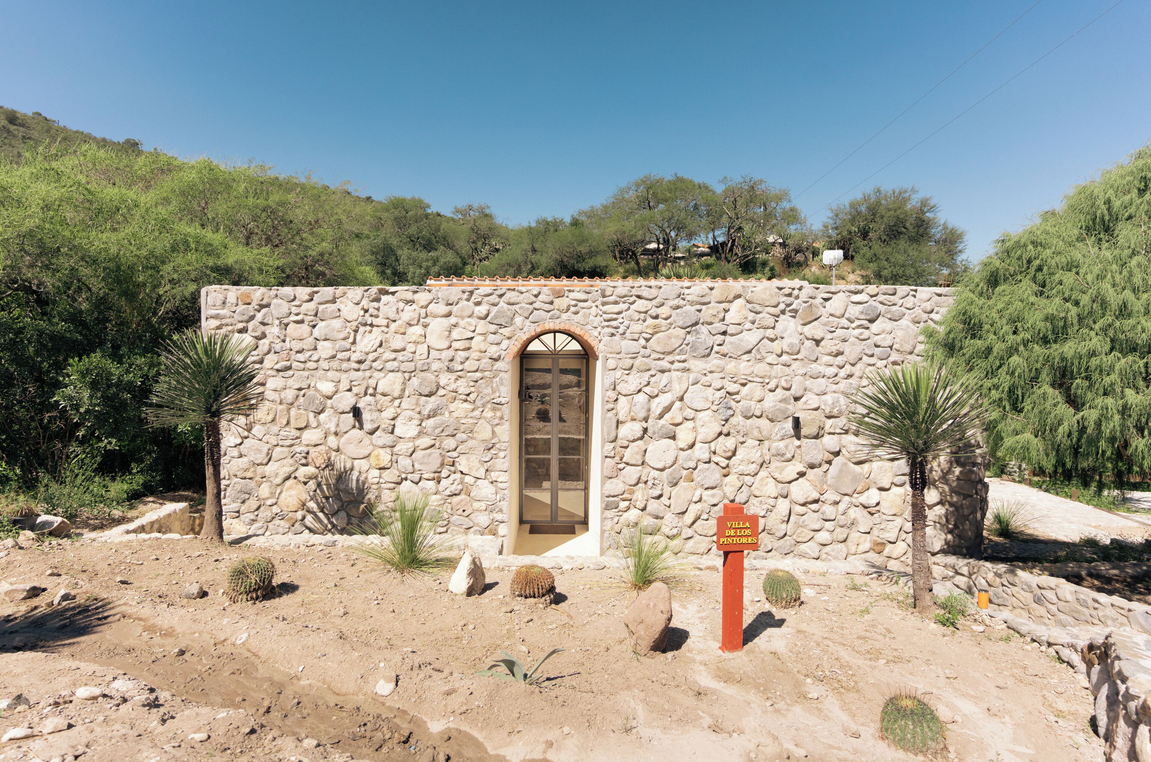 Un bâtiment en pierre doté d’un grand portail en arc de cercle, entouré de cactus et de petits arbres, ce qui suggère un environnement naturel et rustique.