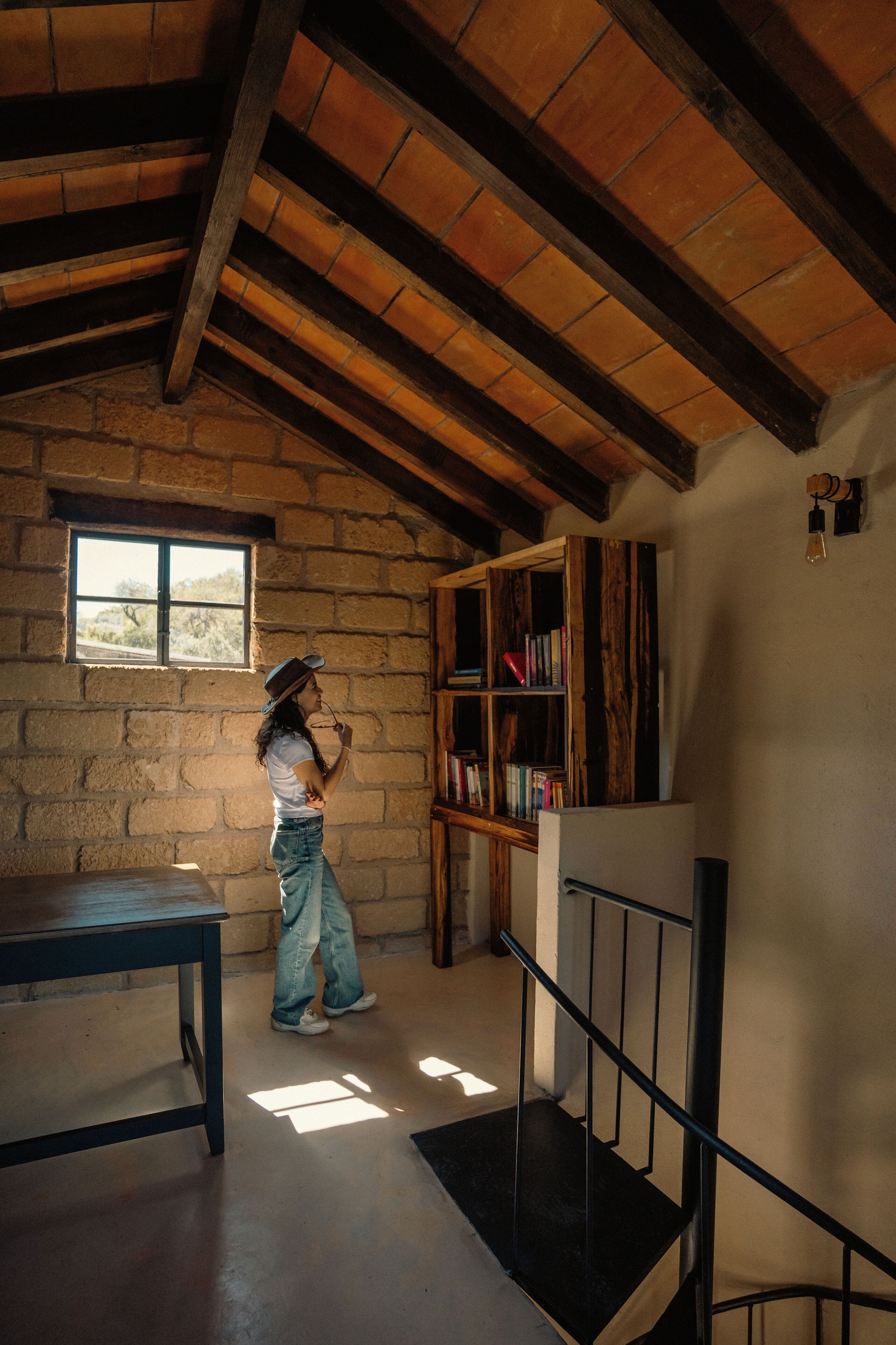 Una habitación rústica con pared de piedra, vigas de madera, una estantería y una ventana.