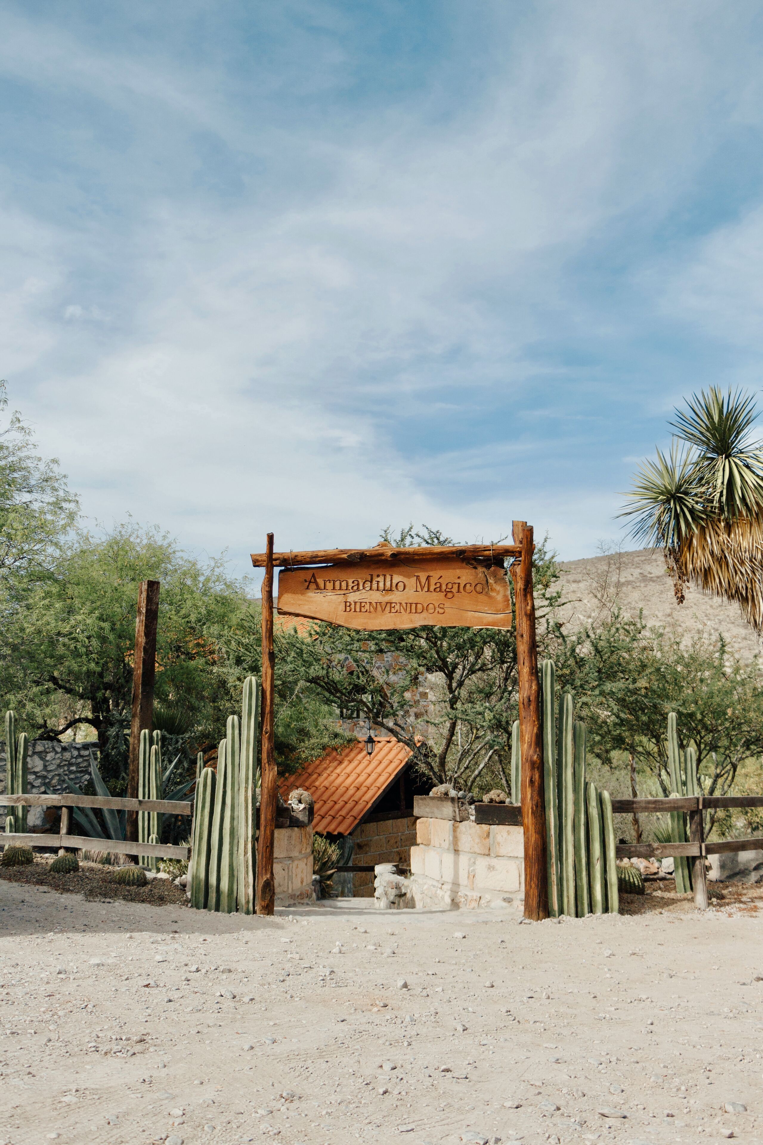 A wooden sign with the text "Armadillo Mágico Bienvenidos" in a desert setting with cacti and a stone building in the background.