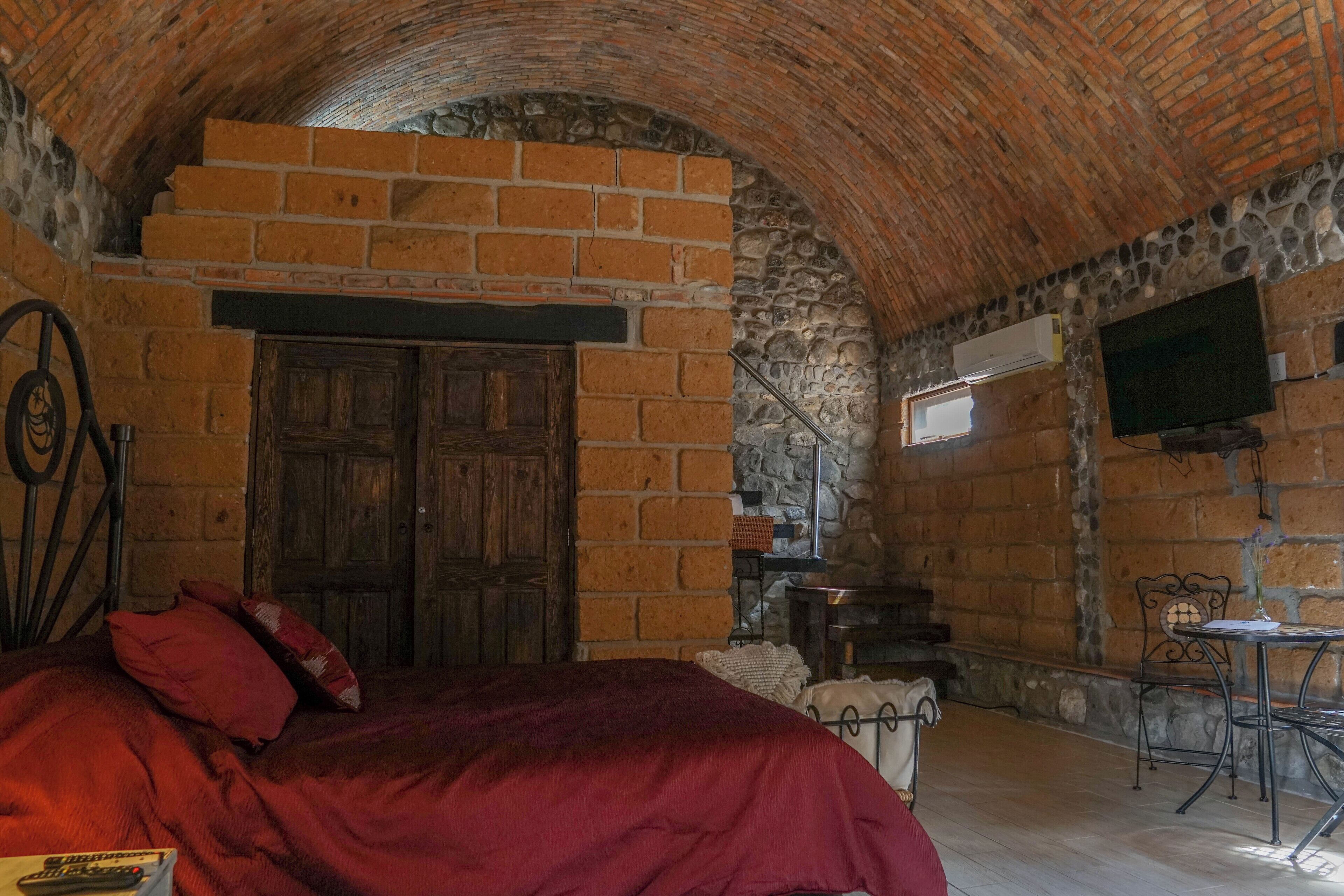 A room with a brick fireplace, a wooden door, a bed with red bedding, a small table, and a television mounted on the wall.