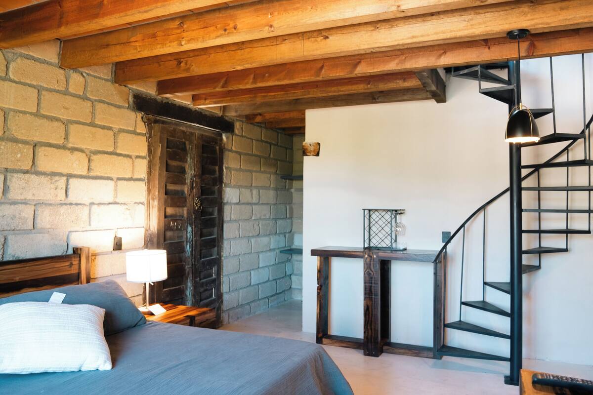 Ein Schlafzimmer mit einem großen Bett, einer Holzbalkendecke, einer Backsteinwand, einer Treppe und einem kleinen Tisch mit einer Lampe.