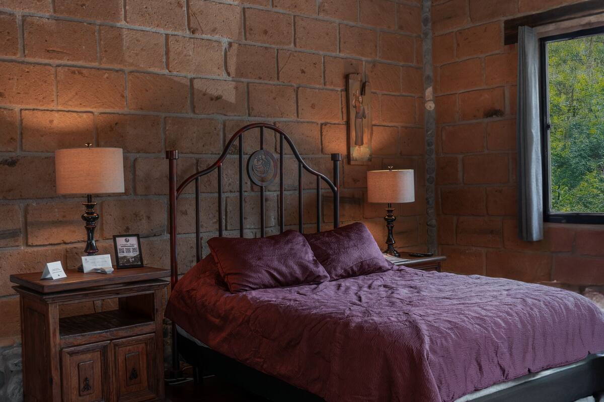 Ein Schlafzimmer mit Backsteinwand, hölzernem Bettgestell und einem Fenster mit Blick auf Bäume.