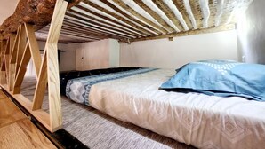 Une chambre située dans un espace de type « loft », avec un plafond en pente, des poutres apparentes, un lit avec une literie à motifs et une fenêtre laissant entrer la lumière naturelle.