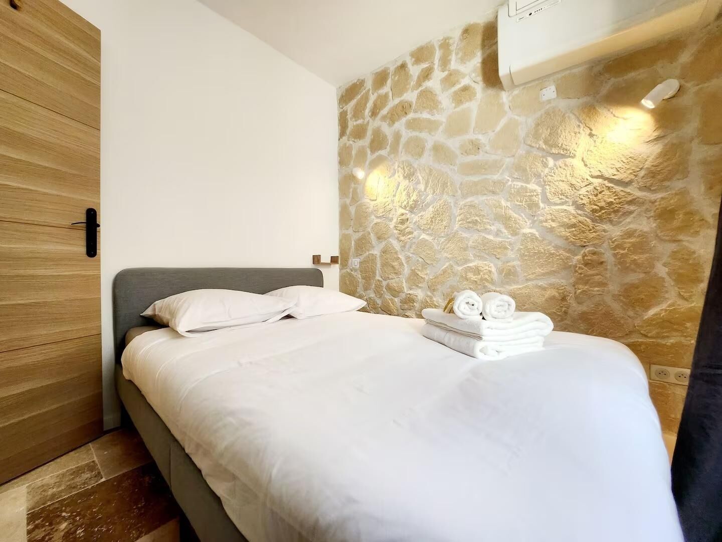 A neatly made bed with white linens and two rolled towels, a stone wall with a warm light, and a wooden door.